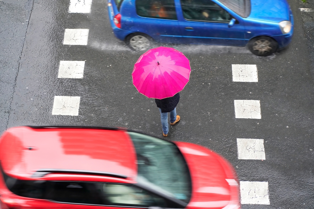 Pedestrian holding a bright pink umbrella standing on a crosswalk while cars drive past on a rainy street.