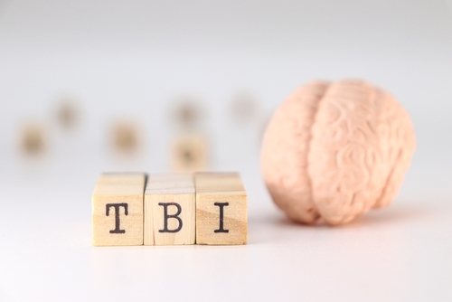 Close up of wooden blocks spelling TBI next to a small model of a human brain on a white background, symbolizing traumatic brain injury awareness.