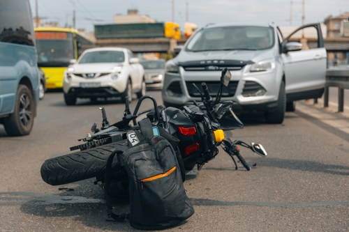 Motorcycle lying on the road after a crash with a silver SUV, backpack nearby, and city traffic in the background.
