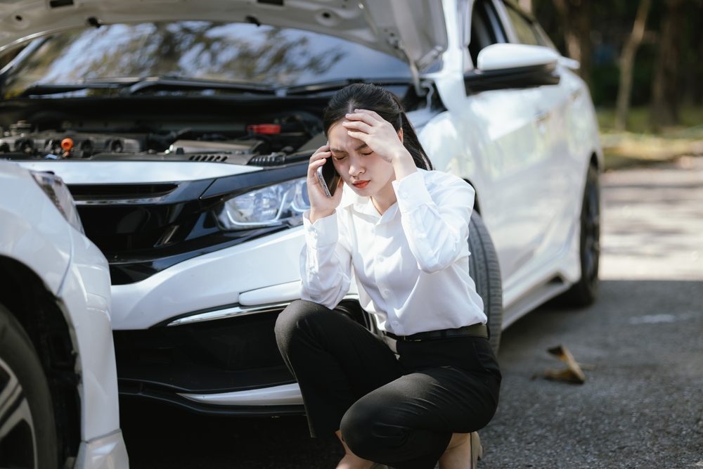 Woman holding her head and calling for help after a car accident with the hood of her car open.