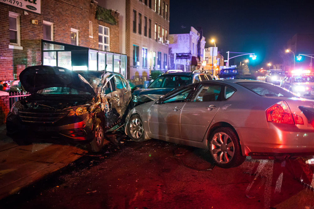 Two heavily damaged cars after a nighttime collision on a city street with emergency lights in the background
