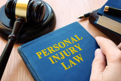 Close-up of a hand resting on a blue law book titled “Personal Injury Law,” with a wooden judge’s gavel placed on the table beside it.