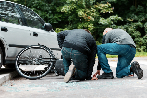 Two men kneeling beside a fallen bicycle and a damaged car after a road accident.