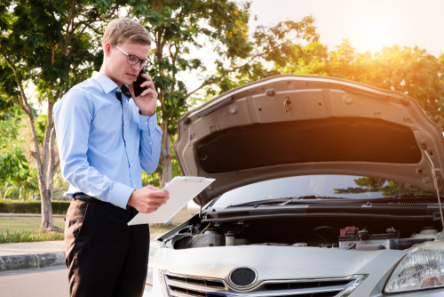 A man wearing a blue shirt and glasses stands beside his car with the hood open, talking on the phone while holding a clipboard under the sunlight.