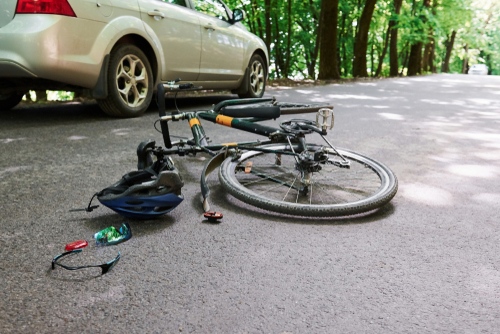 Damaged bicycle lying beside a car on a shaded road after an accident, with a helmet and sunglasses scattered on the ground.