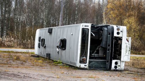 A white bus overturned on its side near a wooded area, showing visible damage to its exterior and windows after a crash.
