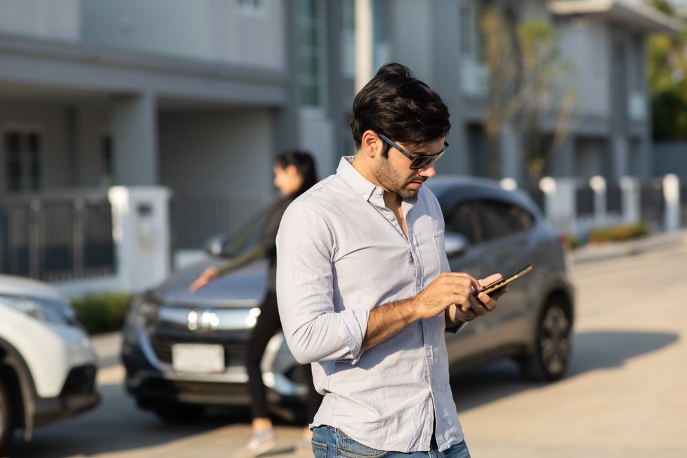 A man wearing sunglasses uses his phone after a minor car accident while a woman stands near the damaged vehicles in the background.