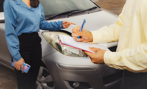 Insurance adjuster writing accident report while woman points at damaged car after collision