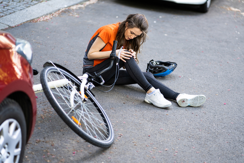 A female cyclist sits on the ground clutching her injured knee next to her fallen bicycle and helmet after a car collision.