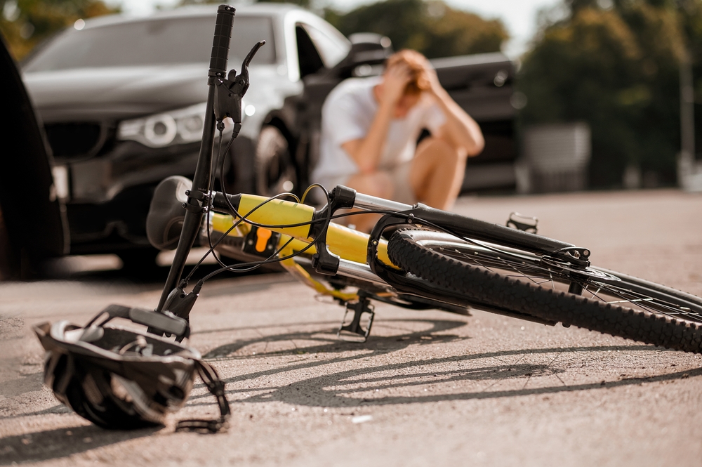 A yellow bicycle lies damaged on the road beside a car, with a distressed cyclist sitting nearby holding his head after an accident.
