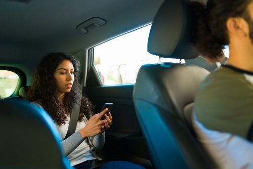 A woman sits in the back seat of a car, wearing a seatbelt and using her phone during a rideshare trip, with the driver visible in front.