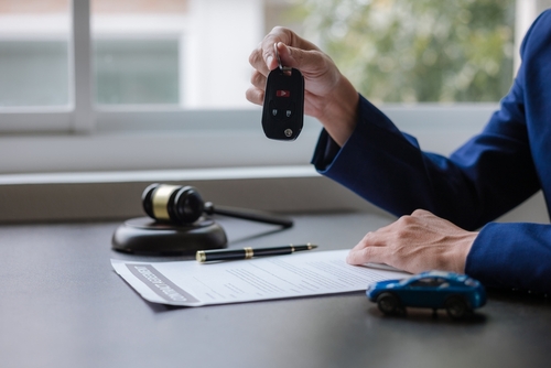 A lawyer holds a car key above a document with a gavel and toy car on the desk, symbolizing car accident claims or vehicle settlements.