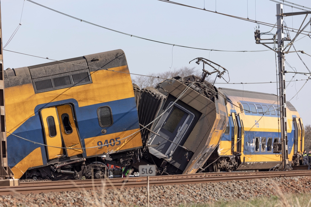 A derailed yellow and blue passenger train lies twisted on the tracks after a major rail accident, with power lines damaged overhead.