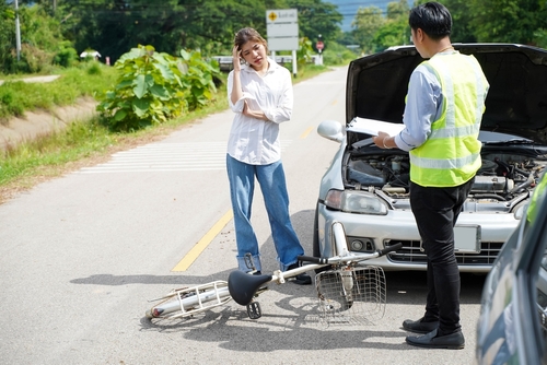 A woman stands beside her damaged car and a fallen bicycle while talking on her phone as a man in a reflective vest inspects the scene of a road accident.