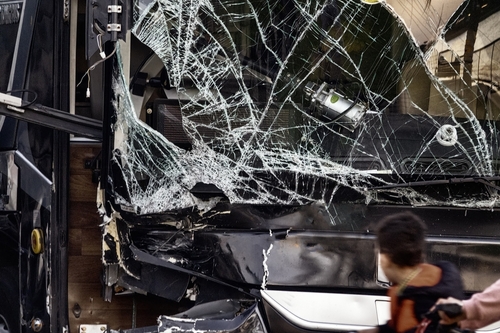 A black bus with a shattered windshield after a serious collision, with a person walking past the damaged vehicle.