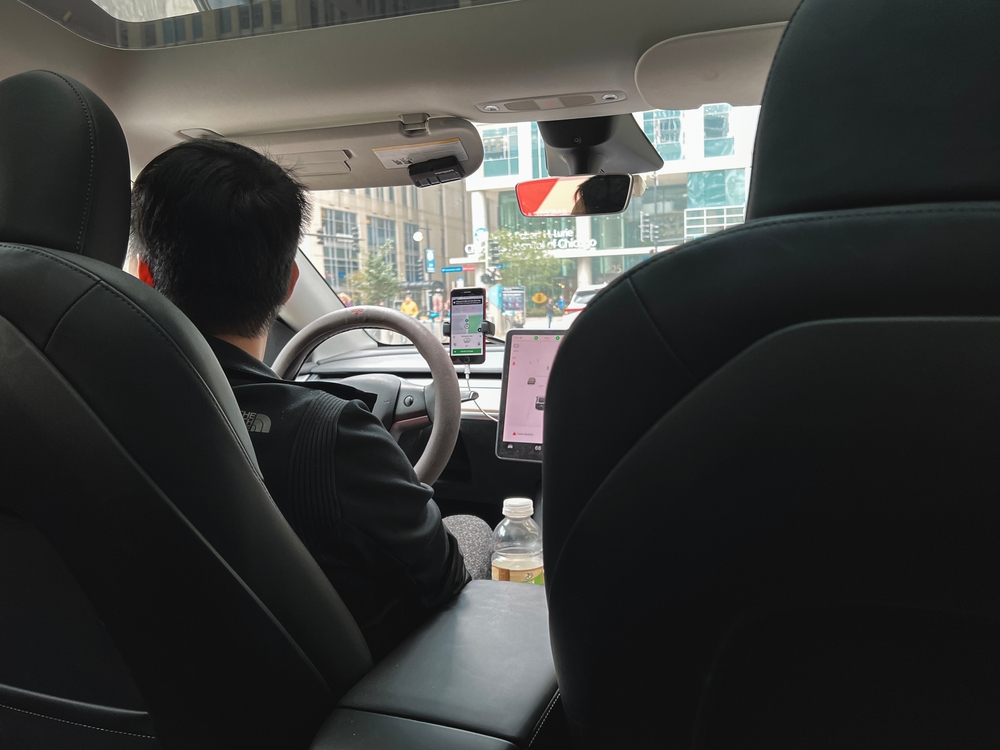 View from the back seat of a rideshare car showing a driver navigating with multiple apps open on mounted smartphones.