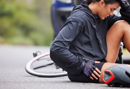A cyclist in a black jacket sits on the road in pain beside her crashed bike after a traffic accident.
