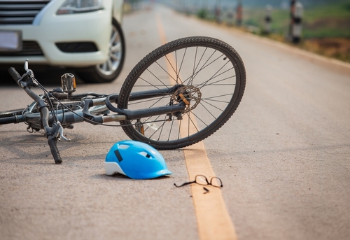 Bicycle lying on the road beside a white car with a blue helmet and glasses scattered nearby.