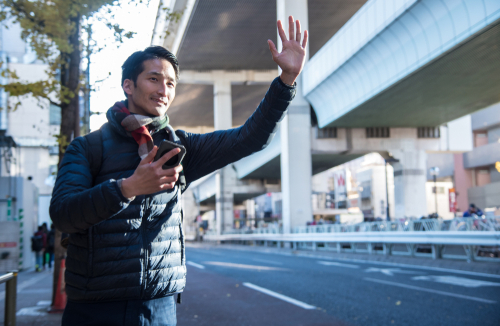 Man standing on a city street holding a phone and raising his hand to hail a rideshare vehicle.